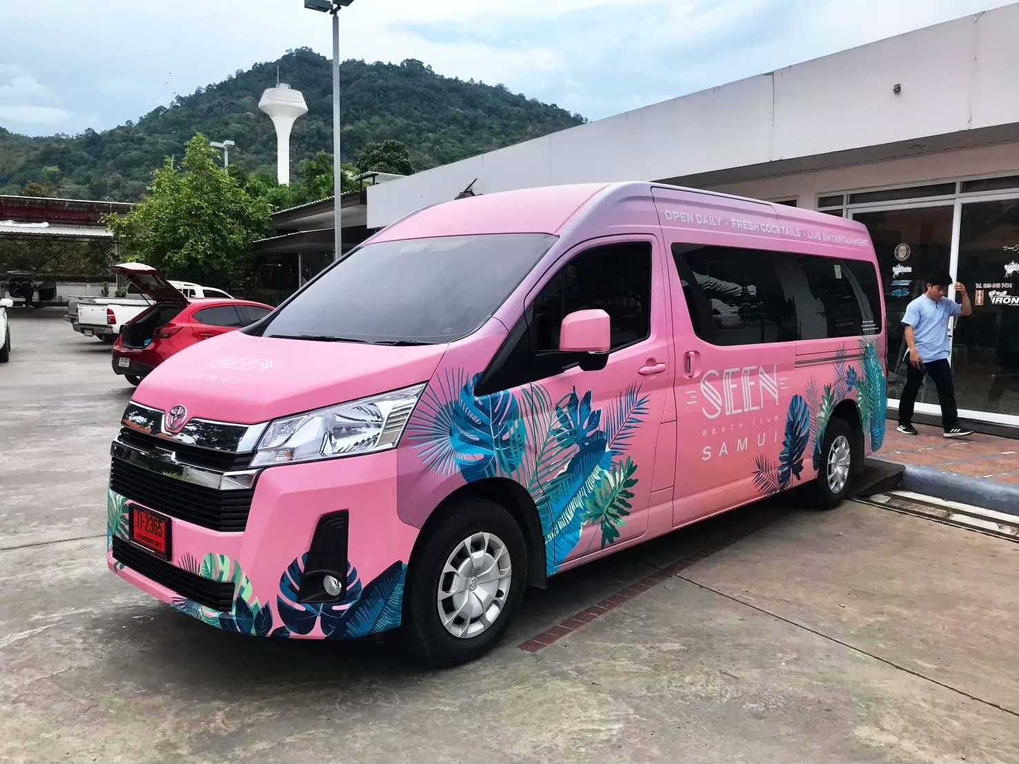 ภาพมุมเฉียงด้านหน้าของรถตู้บริษัท Toyota Commuter แร็ปสติ๊กเกอร์สีชมพูคุมโทนสี CI แบรนด์