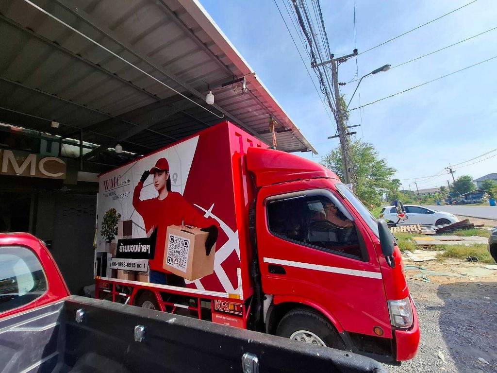 สติ๊กเกอร์แแร๊ปรถขนส่งโลจิสติกส์ ทัศนมิติมุมเฉียงหน้ารถบรรทุก 4 ล้อใหญ่ WMC ย้ายของง่ายๆ โทนสีแดงสดตัดกราฟิกลายเส้นพนักงานหญิงสวมหมวกแก๊ปถือกล่องพัสดุ