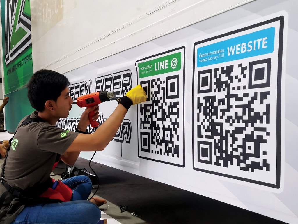 ช่างกำลังใช้ปืนเป่าลมร้อนติดตั้งสติ๊กเกอร์ QR Code และช่องทางติดต่อบนตู้รถ เป็นการอัปเดตข้อมูลตามแผน ดูแลและบำรุงรักษางานฟลีทรถองค์กร
