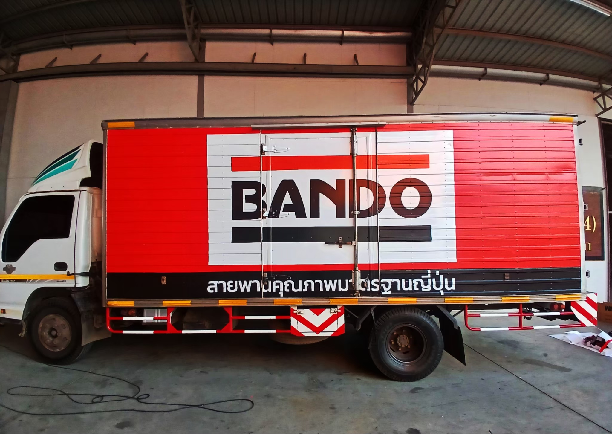 สติ๊กเกอร์รถขนส่งโลจิสติกส์ ภาพถ่ายมุมตรงระนาบข้างฝั่งซ้ายยาวเต็มคันรถบรรทุกหกล้อตู้เหล็ก Isuzu แร๊ปสติ๊กเกอร์สีแดงตัดแบรนดิ้งขาวสลับดำเด่นตาชัดเจน