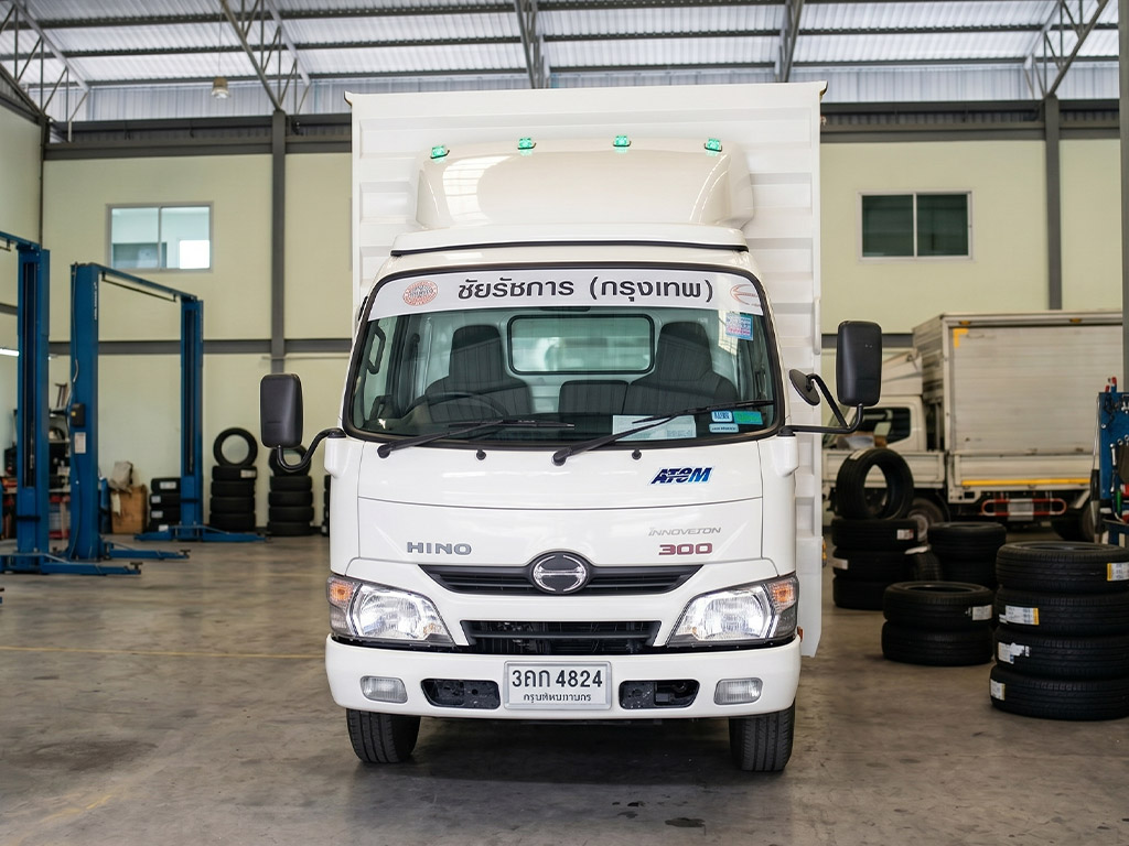 แถบสติ๊กเกอร์หน้ารถขนส่งสินค้าสีขาวพิมพ์อักษรดำบนรถบรรทุก Hino มุมหน้าตรงในโรงปฏิบัติงาน