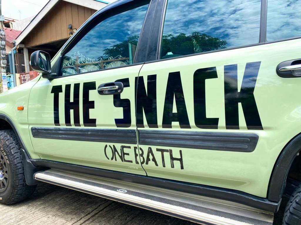 ภาพโคลสอัพตัวอักษรไดคัทสติ๊กเกอร์ชื่อบริษัทบนรถแบรนด์ THE SNACK สีดำแมตต์ จัดวางคร่อมช่องว่างรอยต่อแผงประตูรถด้วยเทคนิคการตัดแบ่งและพับเก็บขอบประณีต