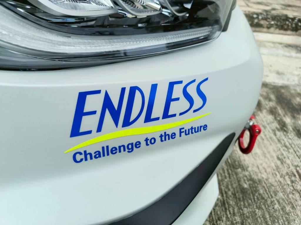 สติ๊กเกอร์ประชาสัมพันธ์เคลื่อนที่ ข้อความ ENDLESS Challenge to the Future สีน้ำเงินเหลืองบริเวณขอบมุมกันชนหน้ารถยนต์สีขาว