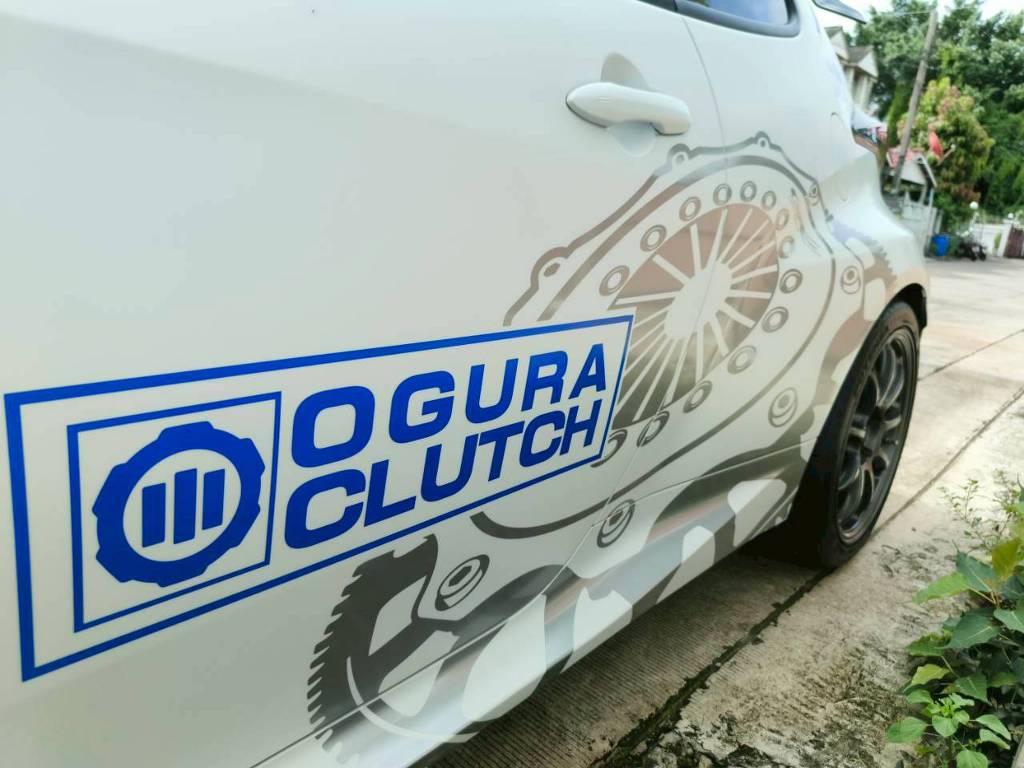 คลังภาพสติ๊กเกอร์ประชาสัมพันธ์เคลื่อนที่ ซูมเจาะลึกโลโก้ OGURA CLUTCH สีน้ำเงินบนพื้นลายกราฟิกกลไกคลัตช์สีเทาข้างประตูรถยนต์