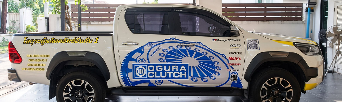 รถกระบะสี่ประตูสีขาวติดตั้งสติ๊กเกอร์ชื่อบริษัทบนรถแบรนด์ OGURA CLUTCH แสดงการจัดวางโลโก้คลัทช์สีน้ำเงินพาดเต็มแนวบานประตูคู่ขนานข้อความภาษาไทยสีเหลืองและคิวอาร์โค้ดสปอนเซอร์