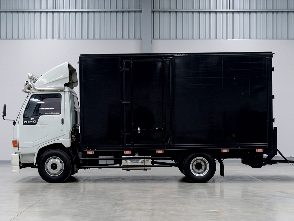รถบรรทุก Hino ตู้ทึบสีดำด้านซ้ายจอดในเวิร์กชอปในร่ม สำหรับเตรียมกระบวนการรื้อสติ๊กเกอร์เก่าและติดตั้งใหม่เพื่อปรับเปลี่ยนโฆษณา