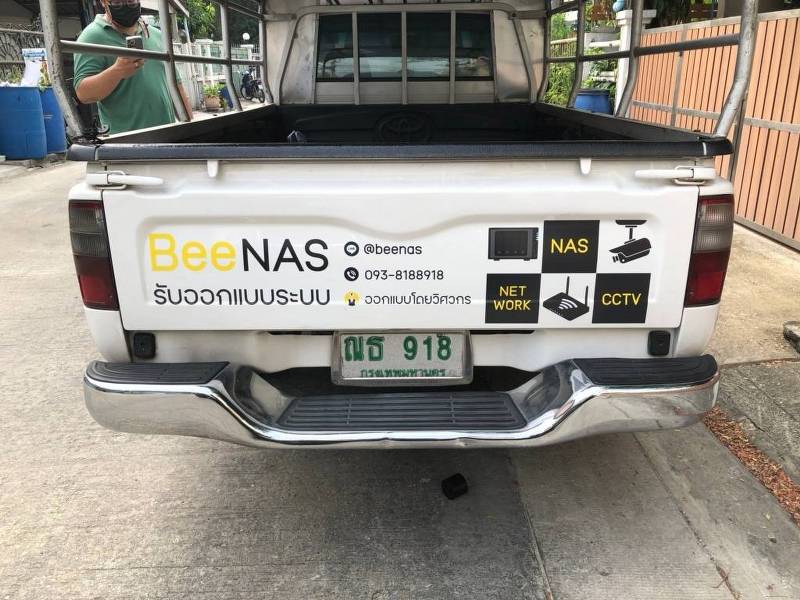 ฝาท้ายรถกระบะติดคอกเหล็กแสดงงานติดตั้งสติ๊กเกอร์ชื่อบริษัทบนรถแบรนด์ BeeNAS พร้อมบล็อกไอคอนสี่เหลี่ยม 2x2 ระบุขอบเขตบริการไอทีชัดเจน