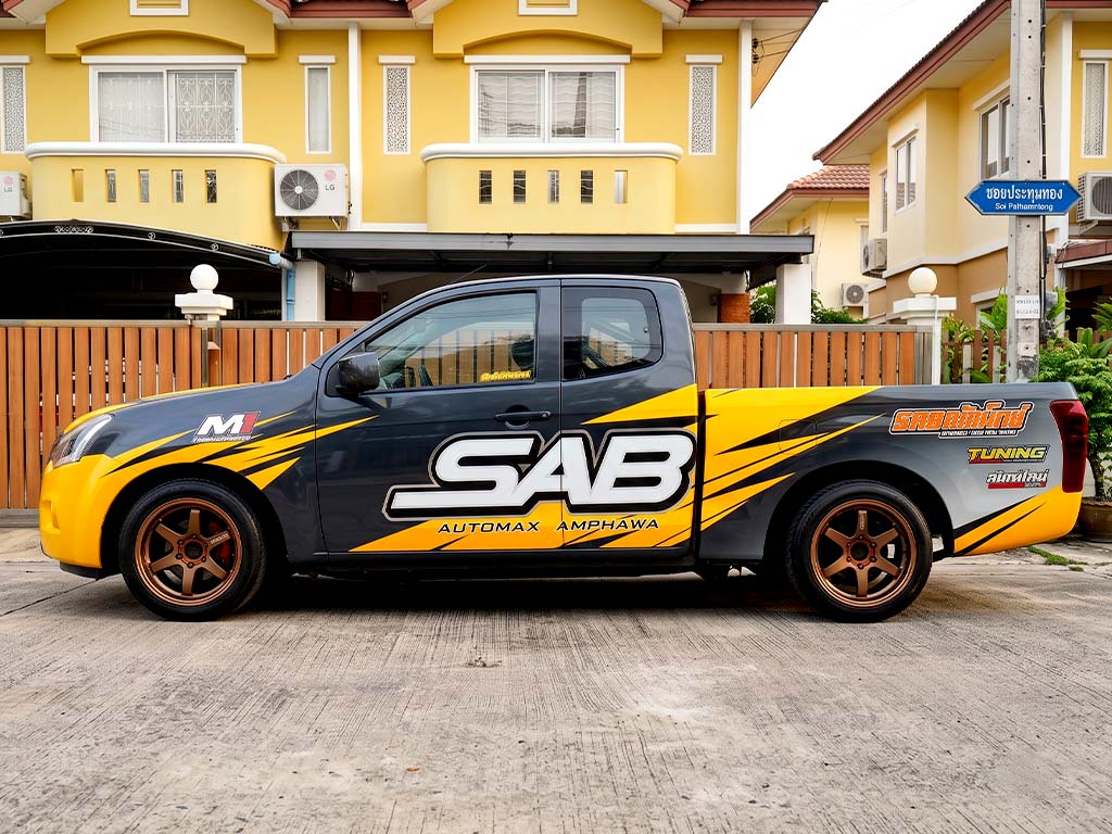 ติ๊กเกอร์รถกิจกรรมและอีเวนต์องค์กร กราฟิกสีเหลืองตัดเทาดำลาย SAB AUTOMAX AMPHAWA บนรถกระบะแคป เน้นความคอนทราสต์สูงช่วยให้อ่านตัวอักษรจากรูปภาพได้ทันที