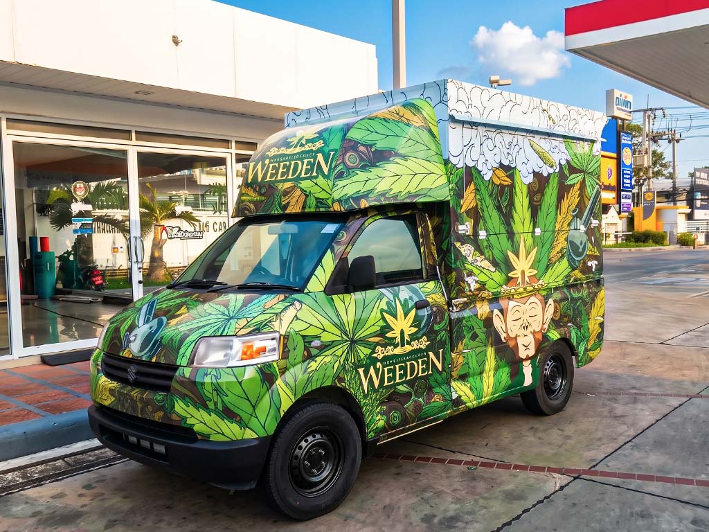 ภาพมุมเฉียงด้านหน้าของรถกระบะ Suzuki Carry หุ้มสติ๊กเกอร์ Full Wrap สีเขียวลายกราฟิกใบไม้และลิงสีน้ำตาล เด่นชัดด้วยโลโก้แบรนด์ WEEDEN เพื่อดึงดูดสายตาในแคมเปญการตลาด