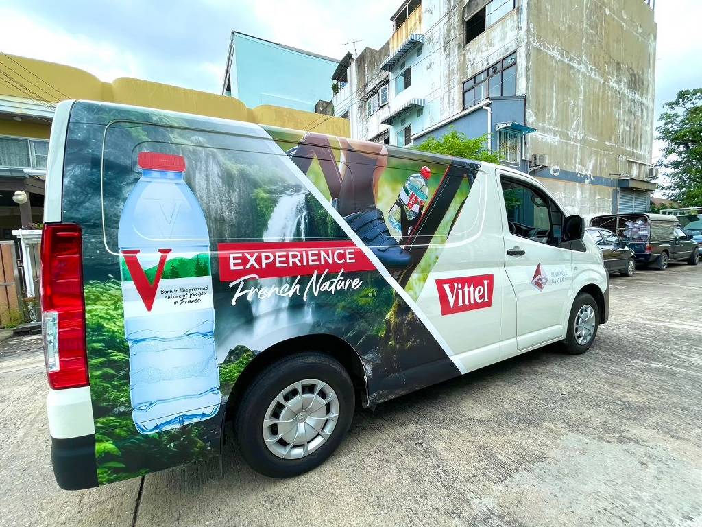 ภาพรวมสติ๊กเกอร์ประชาสัมพันธ์เคลื่อนที่ ลาย Vittel ด้านข้างขวาเต็มคันรถตู้สีขาวท่ามกลางบรรยากาศอาคารพาณิชย์กลางแจ้ง