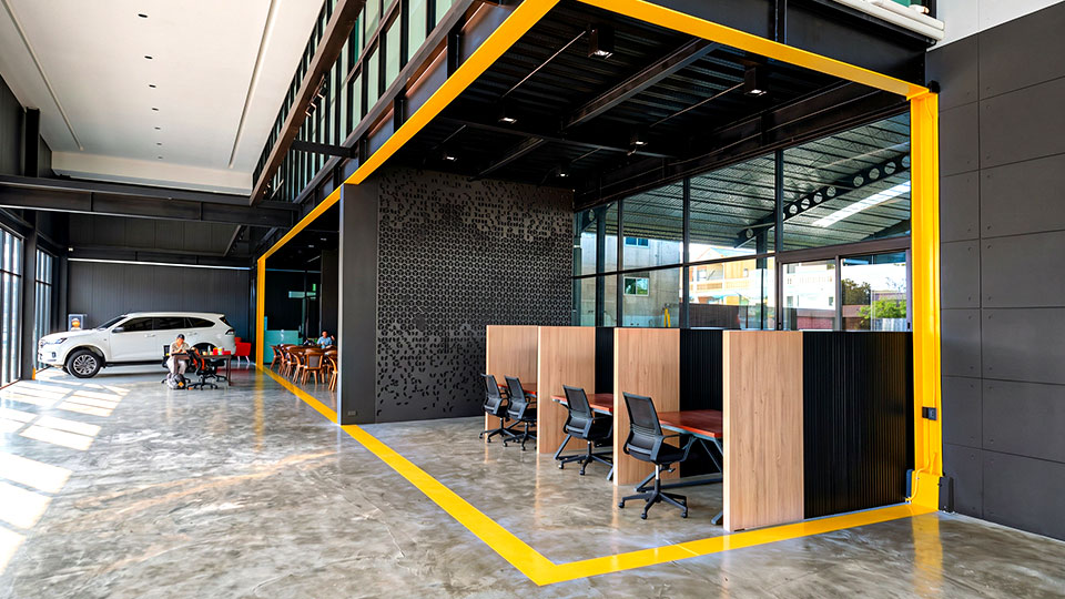 สัญลักษณ์นำสายตาและเส้นแบ่งโซนพื้นที่ (Zoning Graphics) เชื่อมต่อมิติทางสถาปัตยกรรมและนำสายตาลูกค้าอย่างเป็นระบบ การติดตั้งเส้นนำสายตาและกราฟิกแบ่งโซนสีเหลืองบนพื้นคอนกรีตขัดเงา เพื่อจัดระเบียบพื้นและผนังโชว์รูมและสำนักงาน