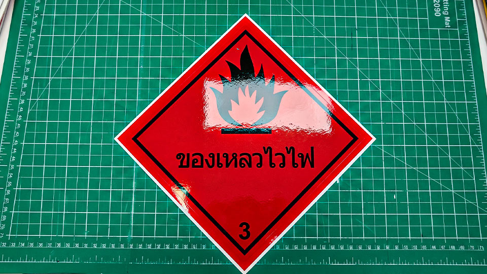 สติ๊กเกอร์เตือนวัตถุอันตราย ของเหลวไวไฟ Class 3 สัญลักษณ์รูปเพชรสีแดงมาตรฐาน GHS วางบนแผ่นตัด