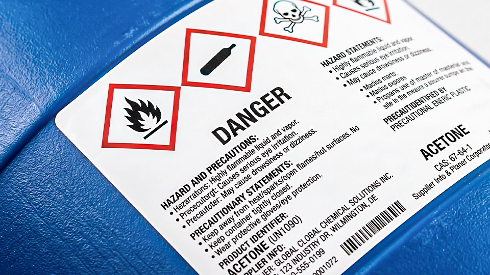 ฉลากสารเคมี GHS อะซิโตน ติดบนถังพลาสติก HDPE สีน้ำเงิน แสดงสัญลักษณ์เตือนเปลวไฟและหัวกะโหลกชัดเจน