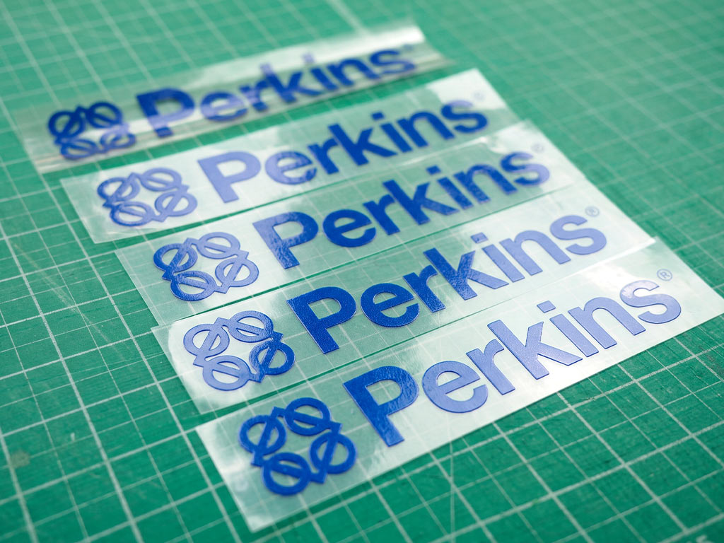 สติ๊กเกอร์ทนความร้อนฟิล์มใส พิมพ์โลโก้ Perkins สีน้ำเงินคมชัด เรียงเป็นชุดบนแผ่นรองตัด