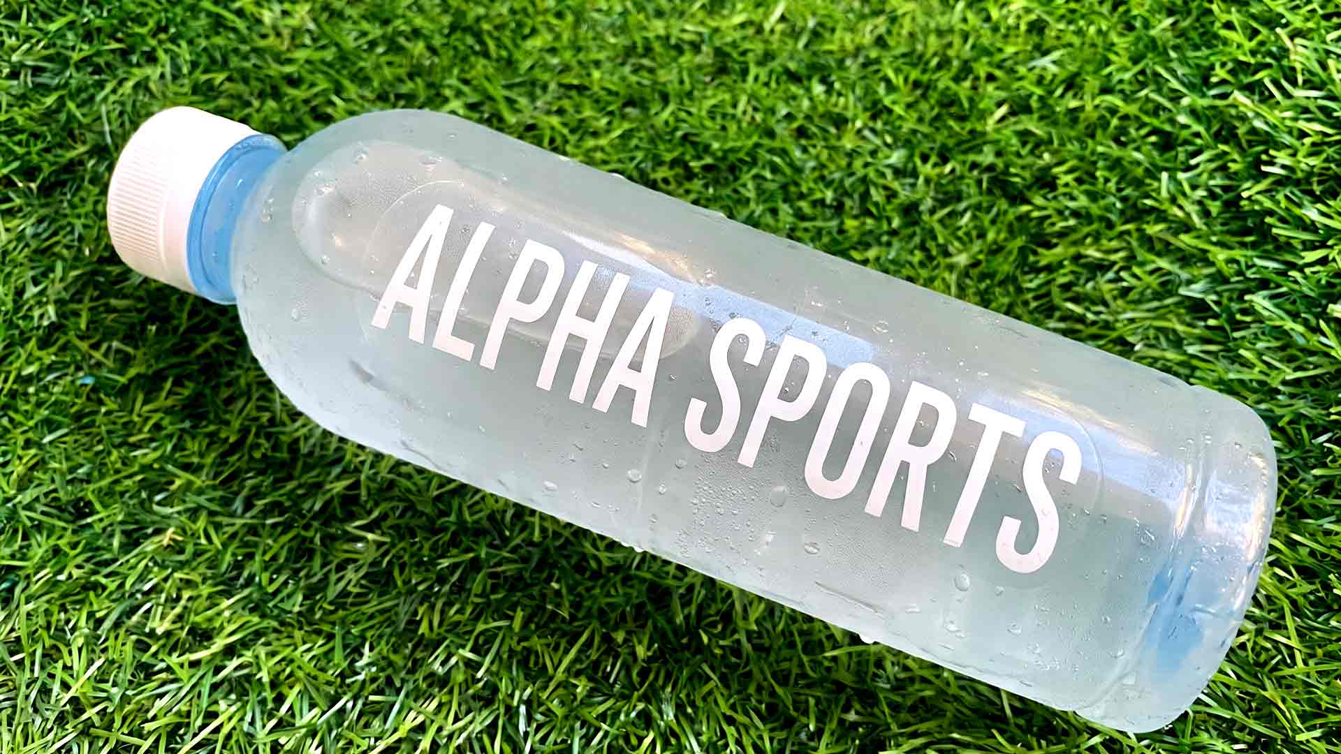 ขวดเครื่องดื่มกีฬา ALPHA SPORTS ติดฉลากใสพิมพ์หมึกขาวทนละอองน้ำและความชื้นจากการแช่เย็น วางบนพื้นหญ้าเทียม