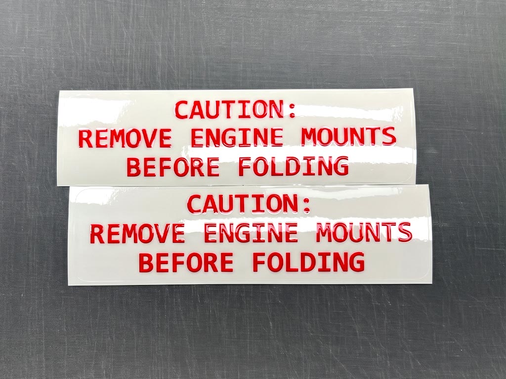 ป้ายความปลอดภัยโรงงาน มุมมองตรงของสติ๊กเกอร์ CAUTION REMOVE ENGINE MOUNTS BEFORE FOLDING 2 ชิ้น