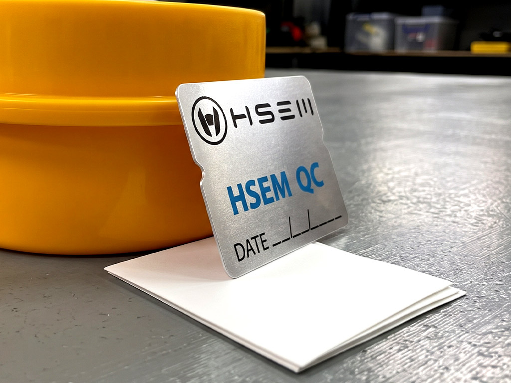 ฉลาก QC, Lot, Batch และ Traceability สติ๊กเกอร์ฟอยล์เงิน HSEM QC มุมมองเฉียงขวาเผยขอบไดคัทรอยบากเว้าคมกริบ